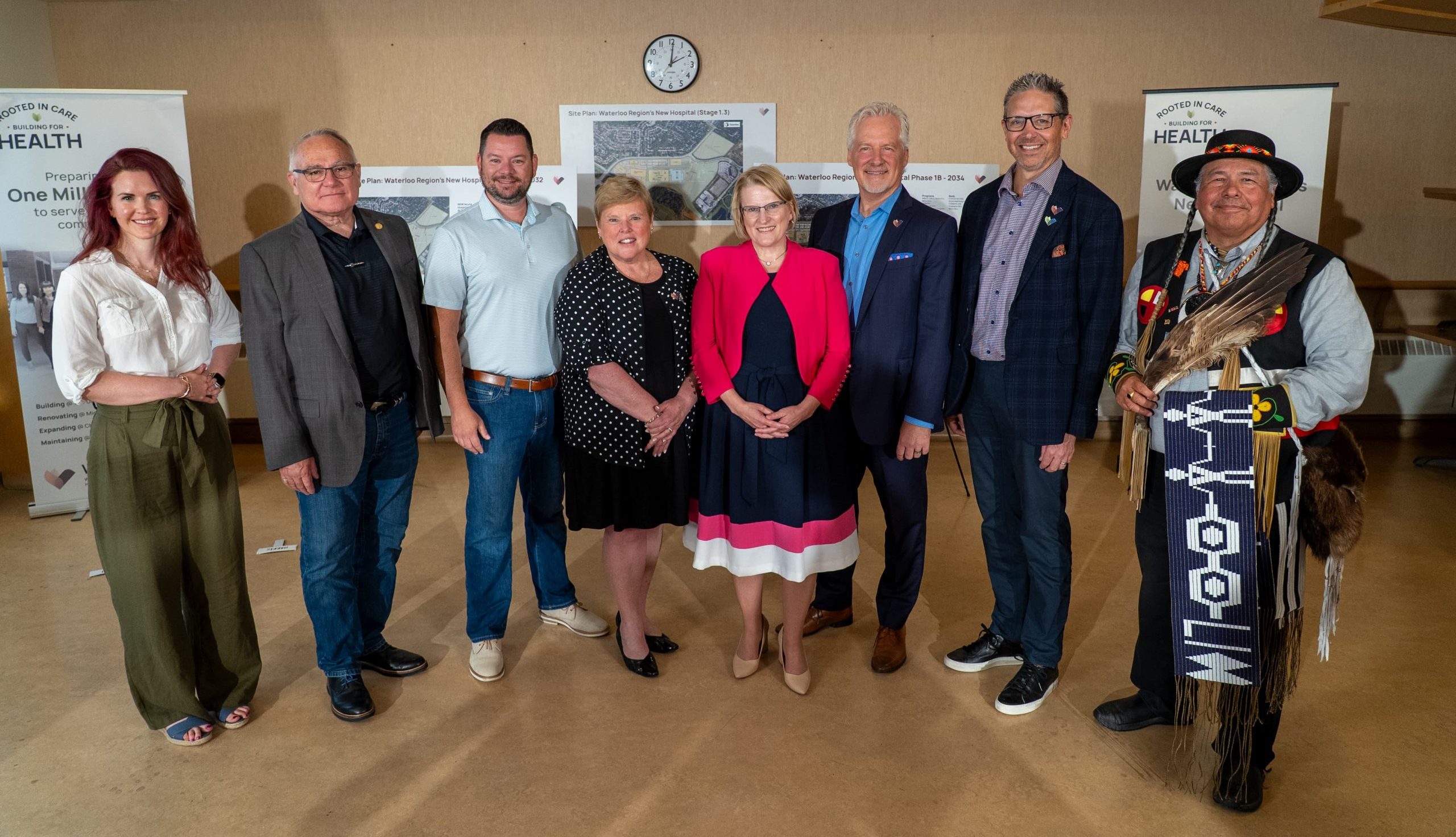 Eight people stand indoors in a row, posing for a group photo in front of informational banners and a clock.
