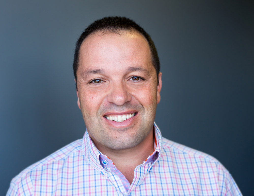 Angelo in a checked shirt standing in front of a plain dark background.