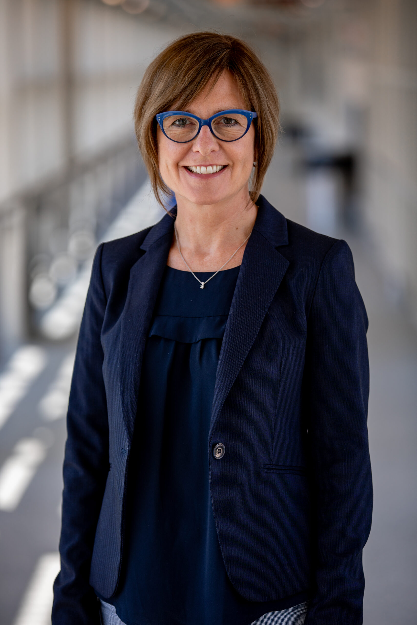 A woman with short brown hair and glasses smiles, wearing a black top and standing indoors.