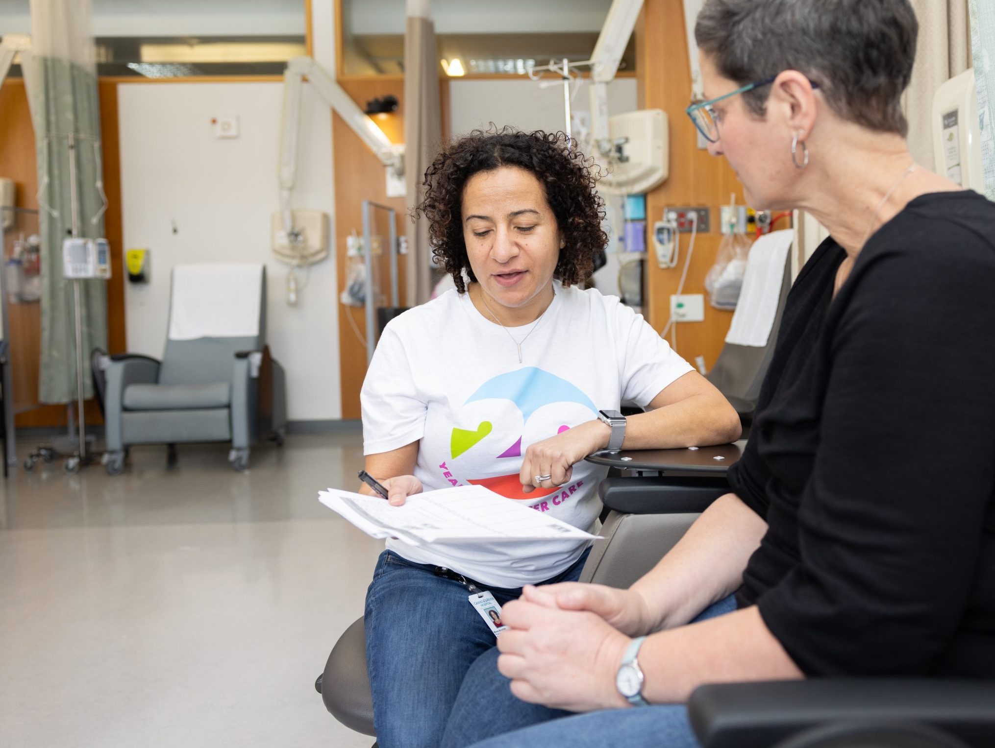 Two women sit in a medical clinic; one reviews paperwork while the other listens attentively.
