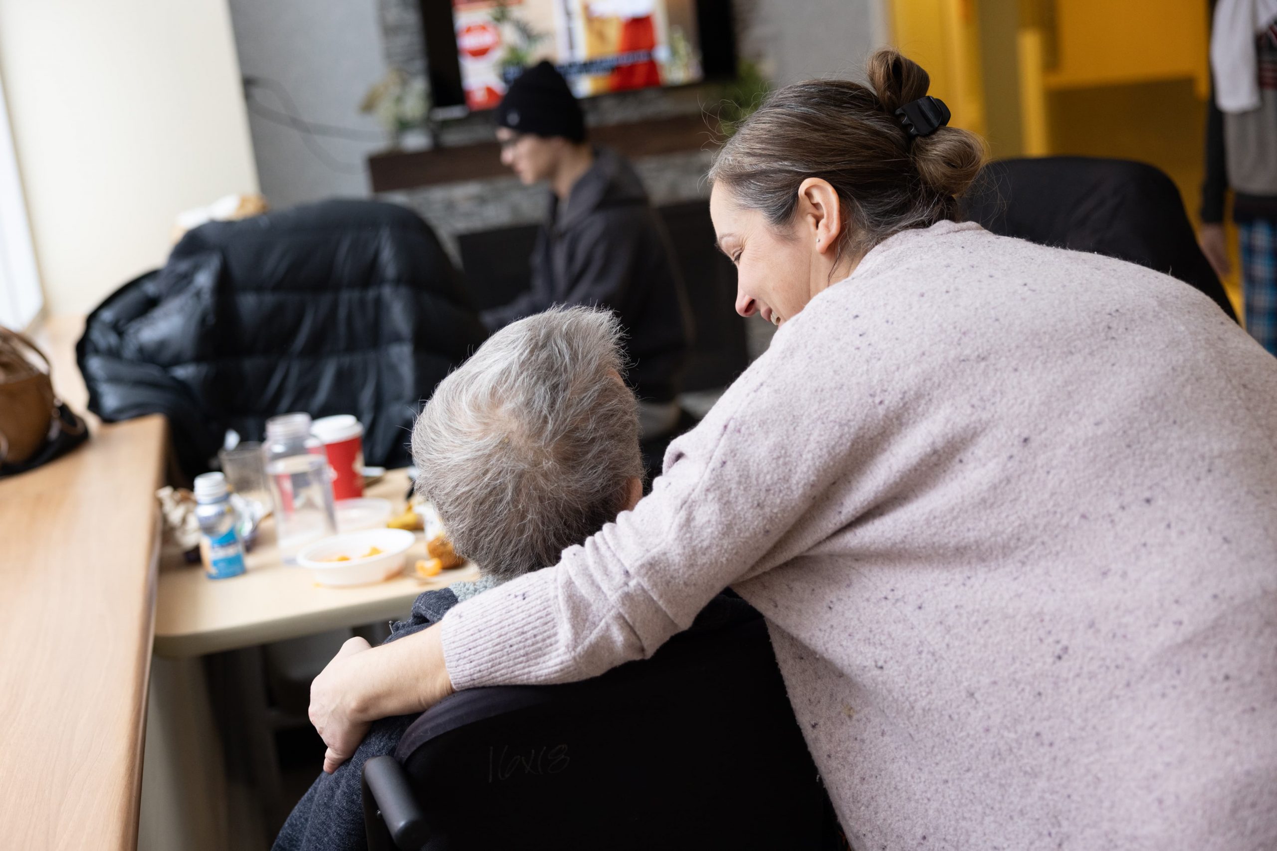 A woman leans over to embrace an elderly person in a wheelchair near a table with food and drinks.