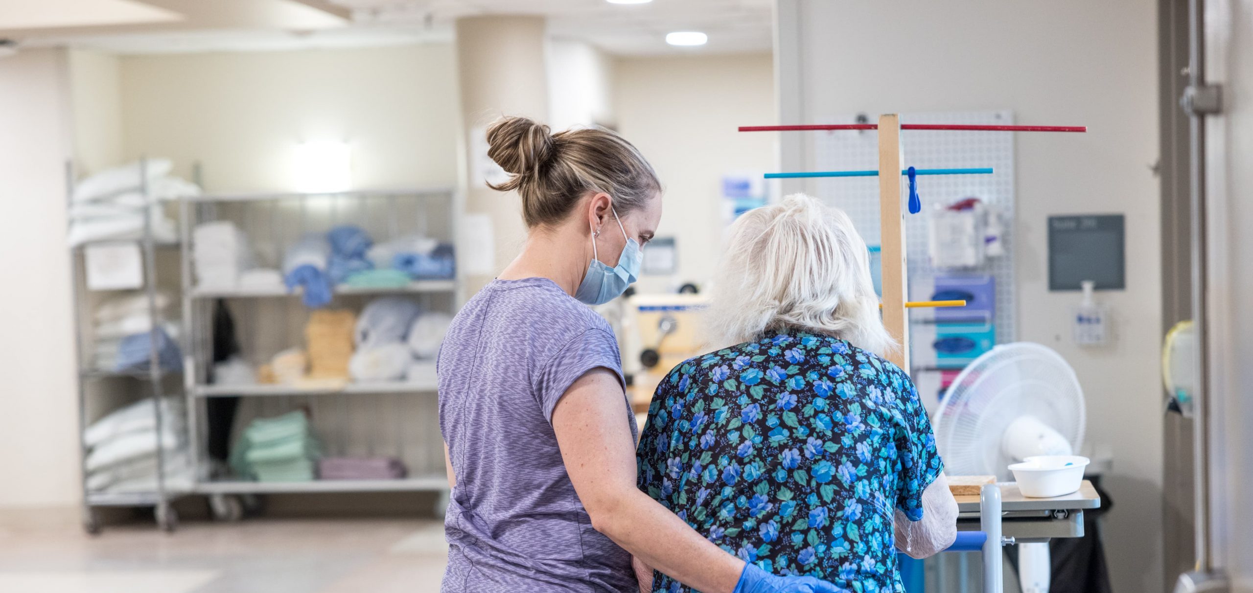 A healthcare worker assists an elderly woman walking down a hallway in a medical facility.