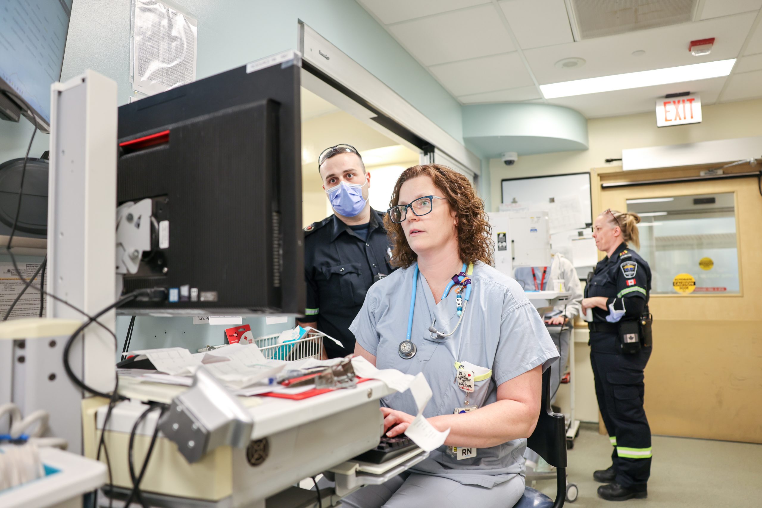 A nurse works at a computer station while two paramedics, one masked, stand nearby in a hospital setting.