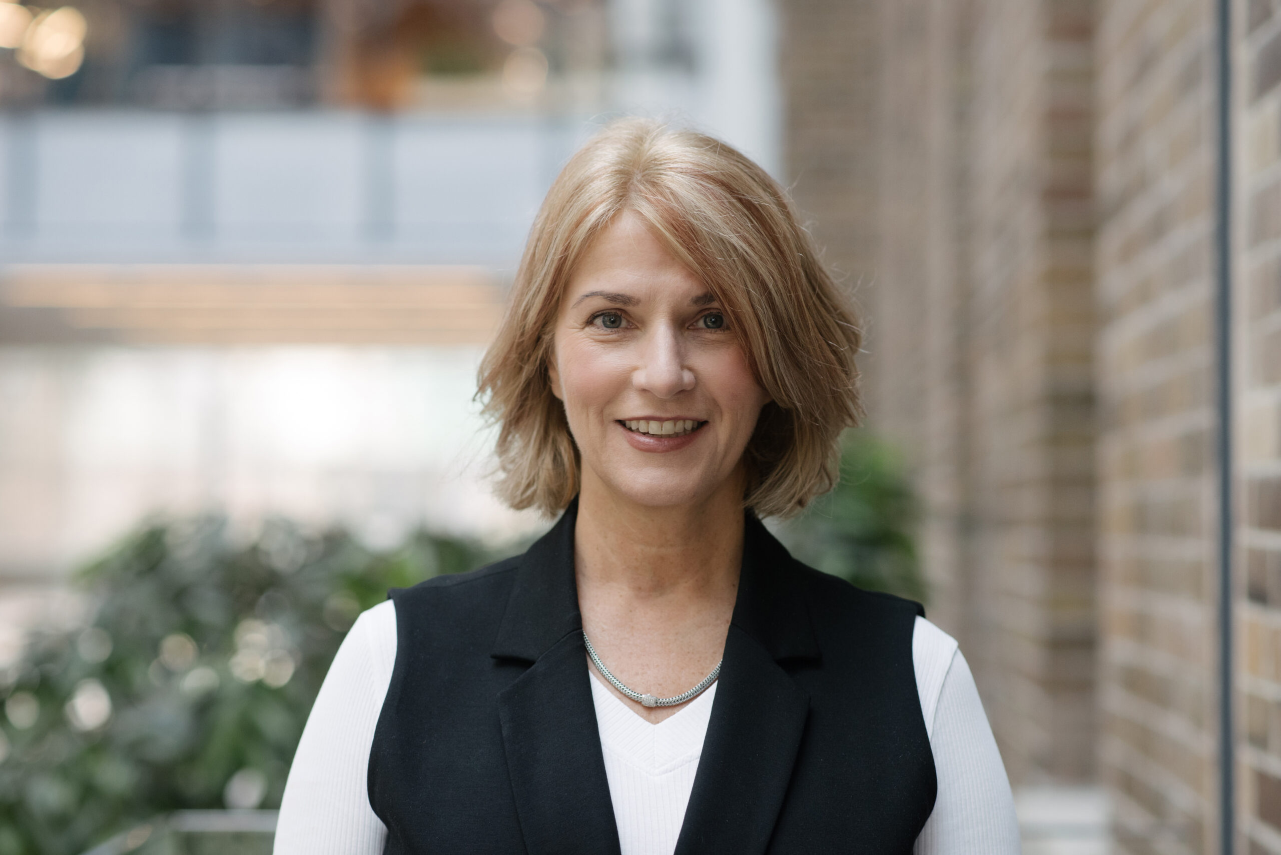 Stephanie with short blonde hair, wearing a black vest over a white top, stands indoors in front of a blurred background.