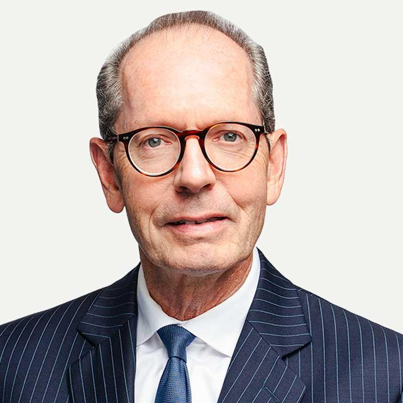 Tom poses in a pinstripe suit, glasses, and a blue tie, facing forward against a plain light background.