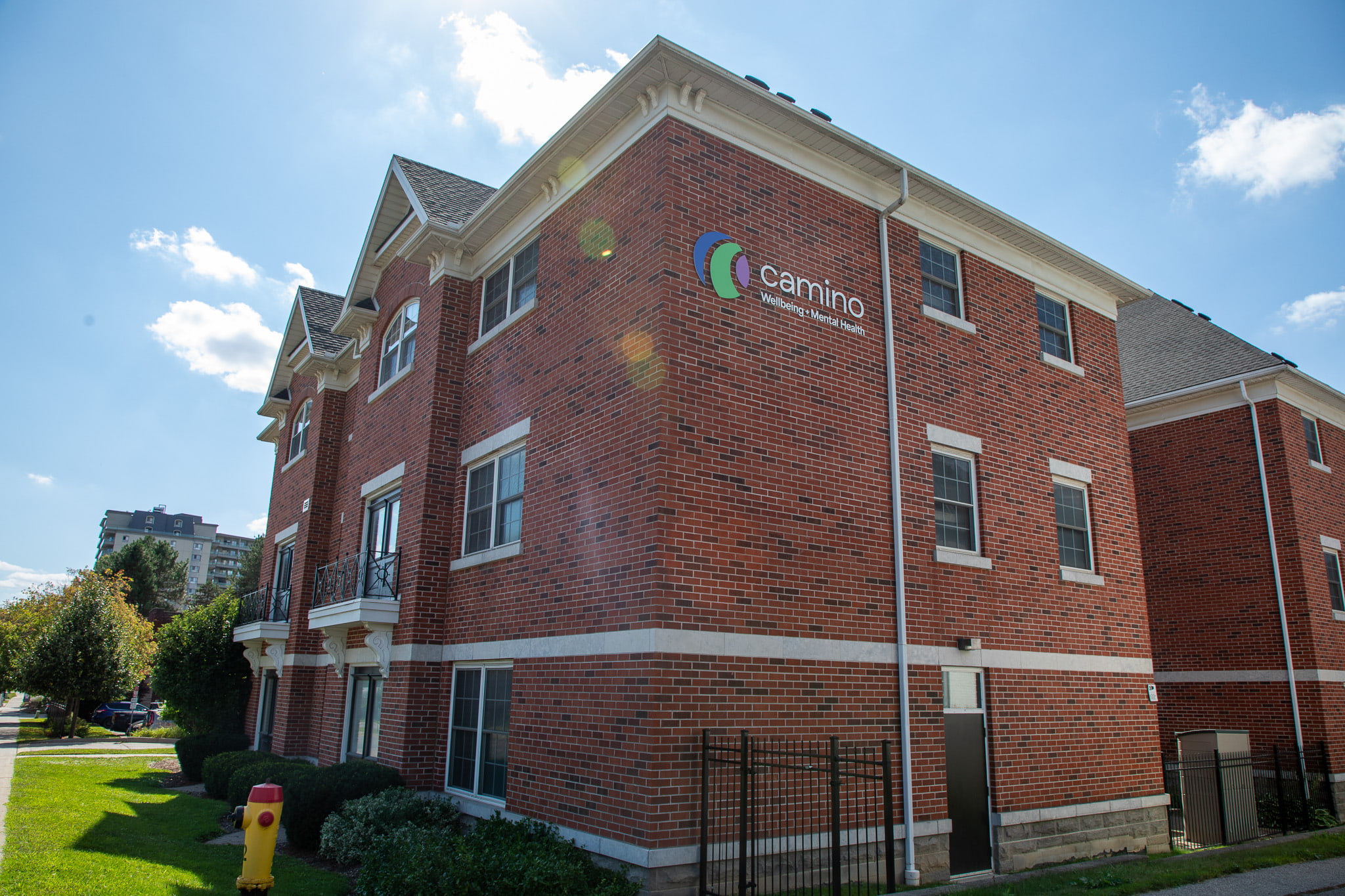 Three-story red brick building with Camino sign on the side, surrounded by grass and shrubs on a sunny day.