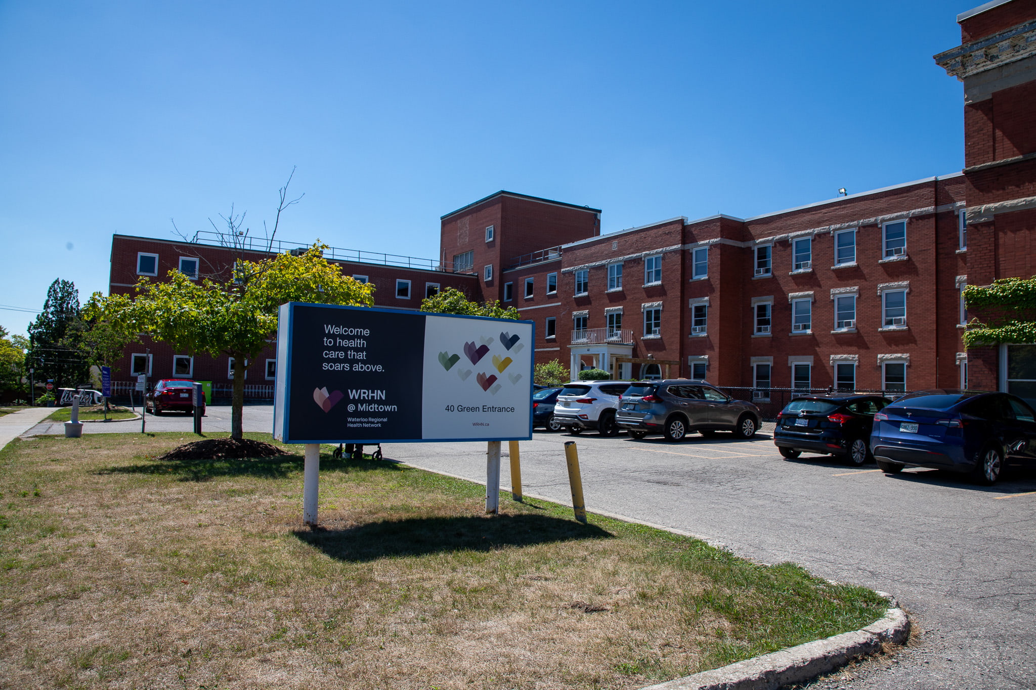 A brick healthcare facility with a parking lot and a welcome sign in front on a sunny day.