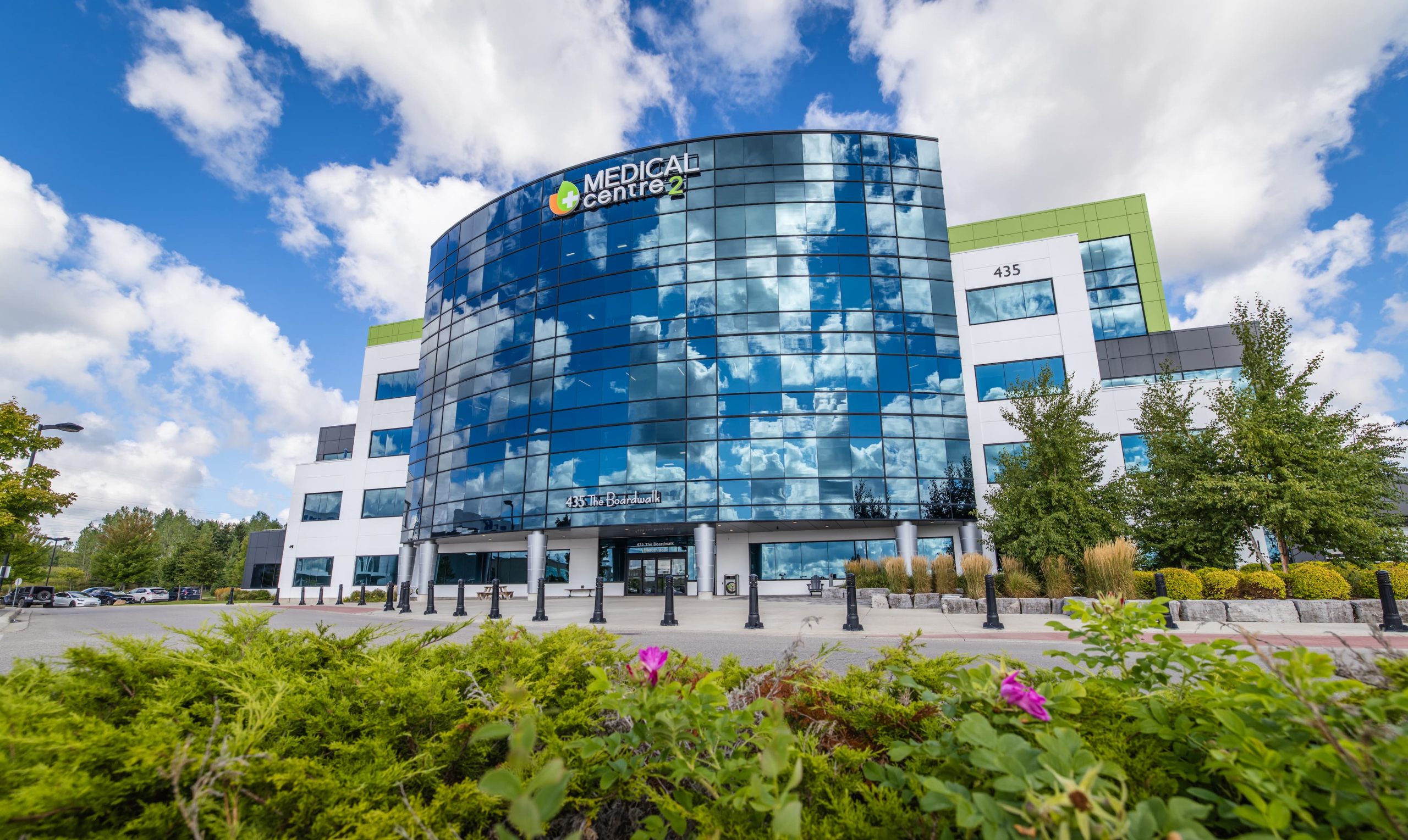 Modern glass-fronted medical center building with clouds reflected on windows, surrounded by greenery and flowers.