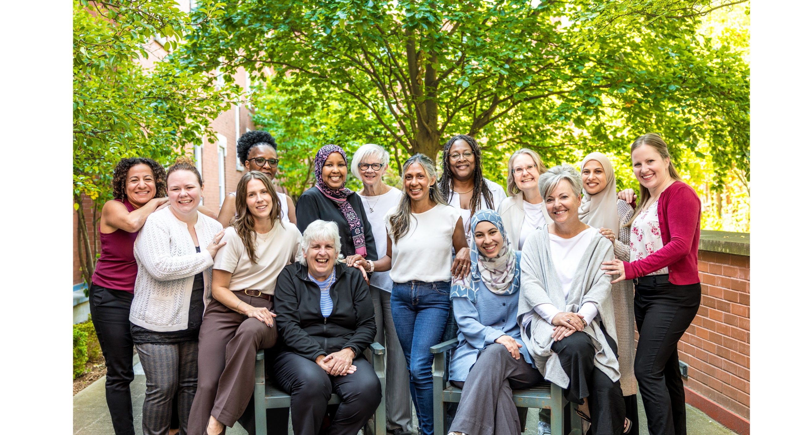 十五名女性在树木与砖墙前户外合影，她们共同摆出姿势并展露笑容。