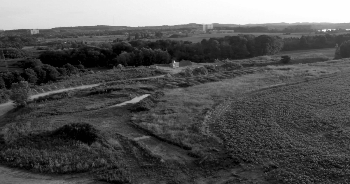 Aerial view of a rural landscape with fields, a dirt path, trees, and distant buildings on the horizon in black and white.