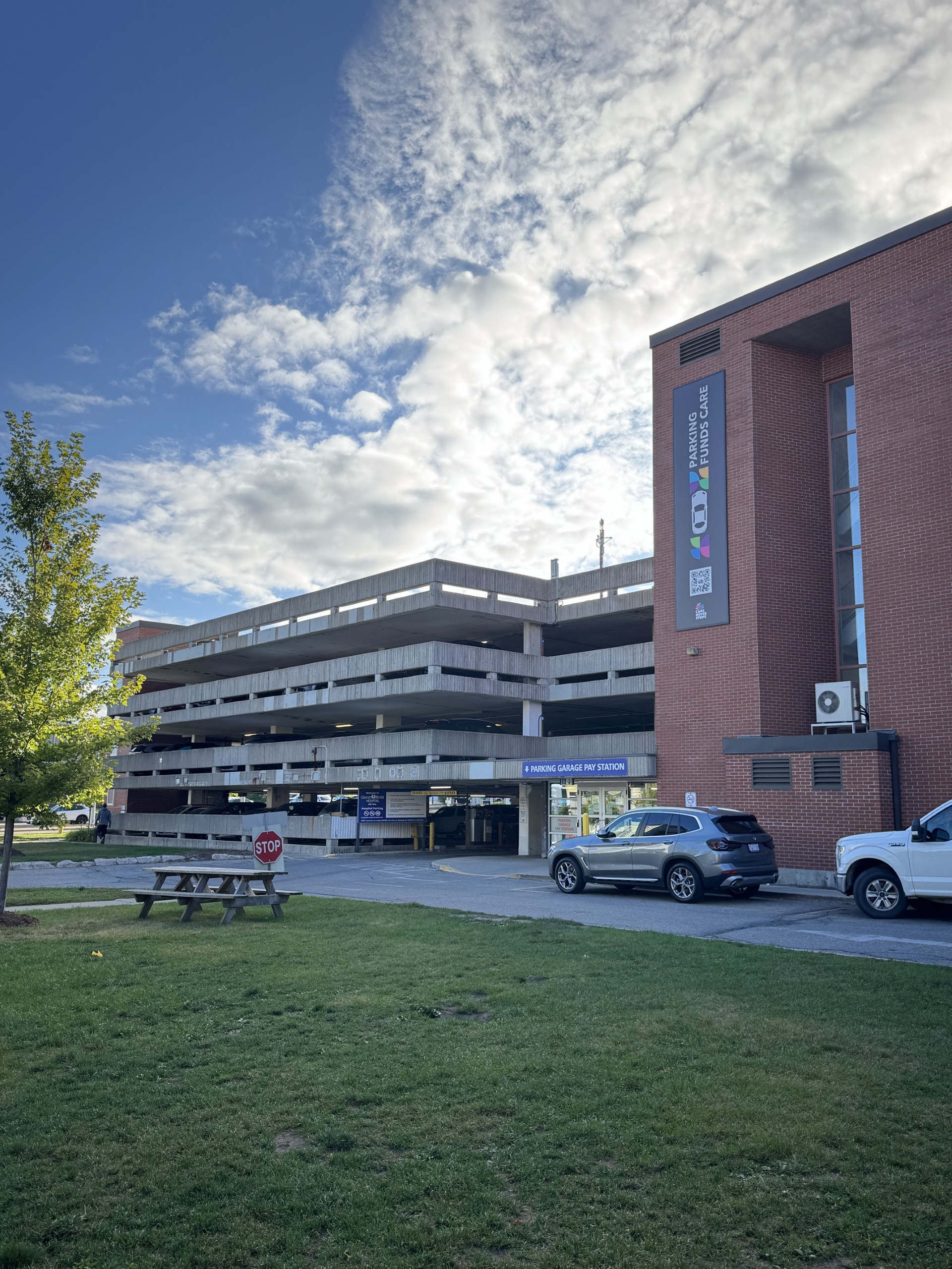A multi-level parking garage and a brick building under a partly cloudy sky, with cars parked and a picnic table nearby.