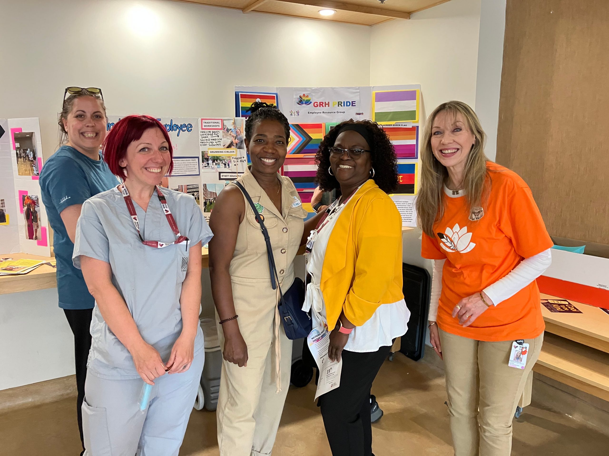 Five women stand and smile in front of a display board featuring various posters and rainbow flags in a brightly lit room.