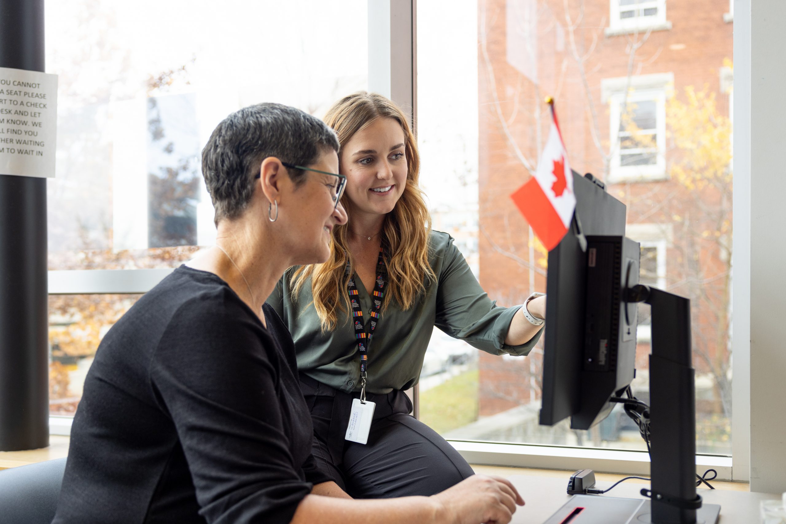 Two women sit at a desk, looking at a computer monitor with a small Canadian flag attached, in an office setting.