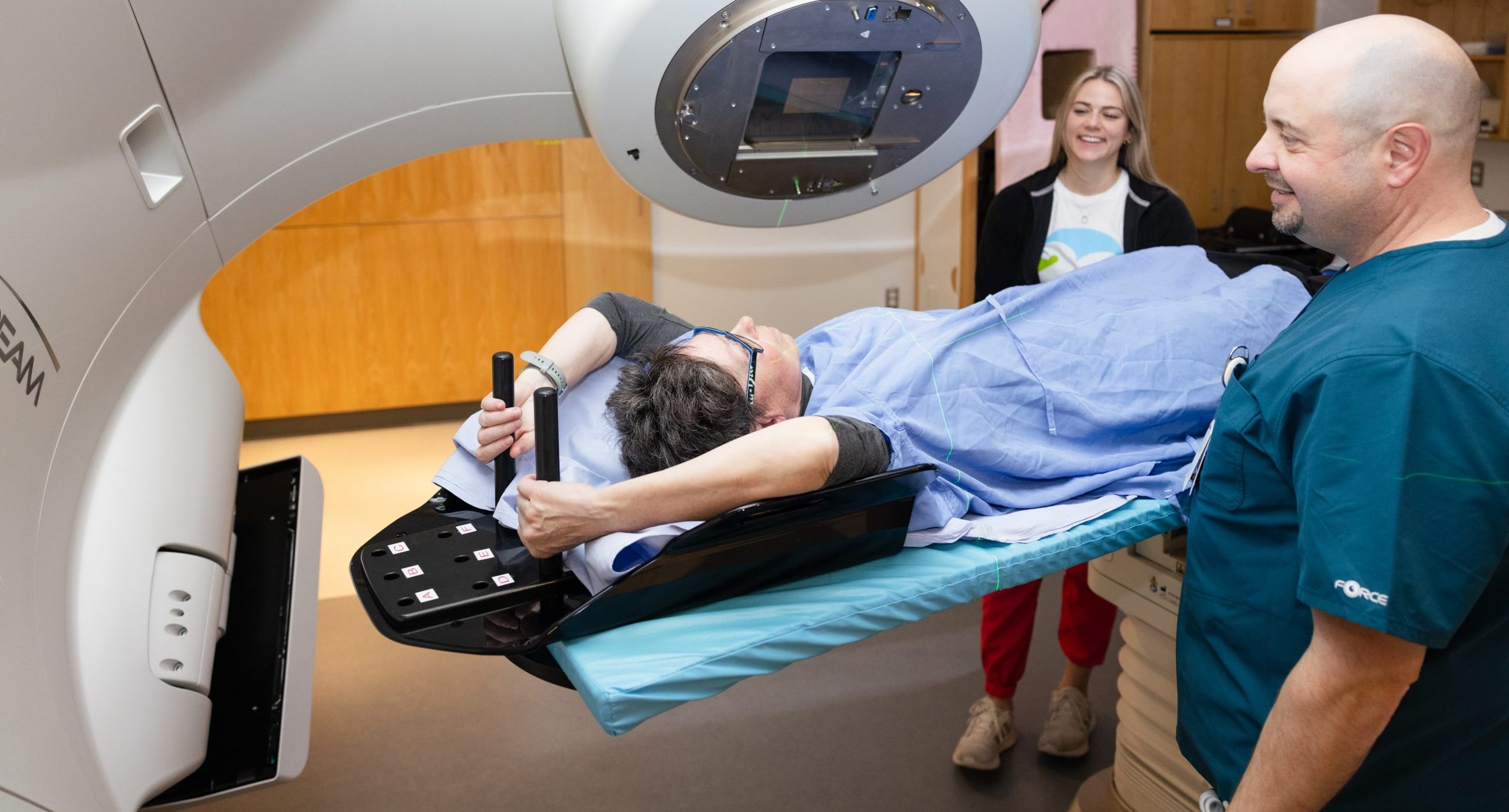 A patient receives radiation therapy while lying on a treatment table, monitored by two medical staff in a clinic.