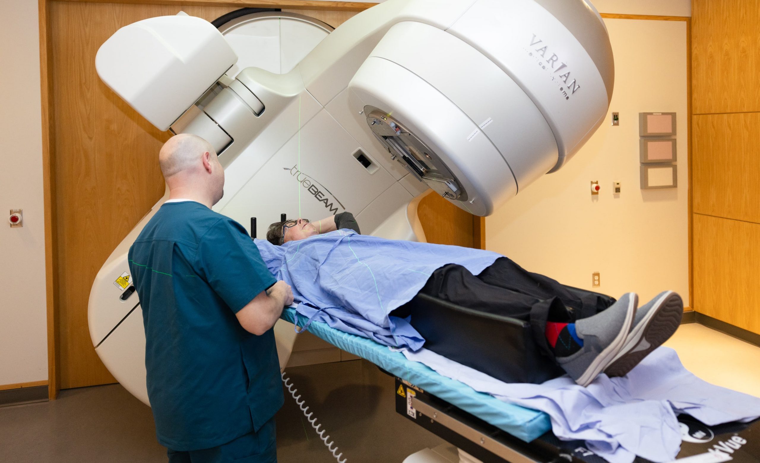 A patient lies on a table under a large Varian medical machine while a healthcare worker stands beside them.