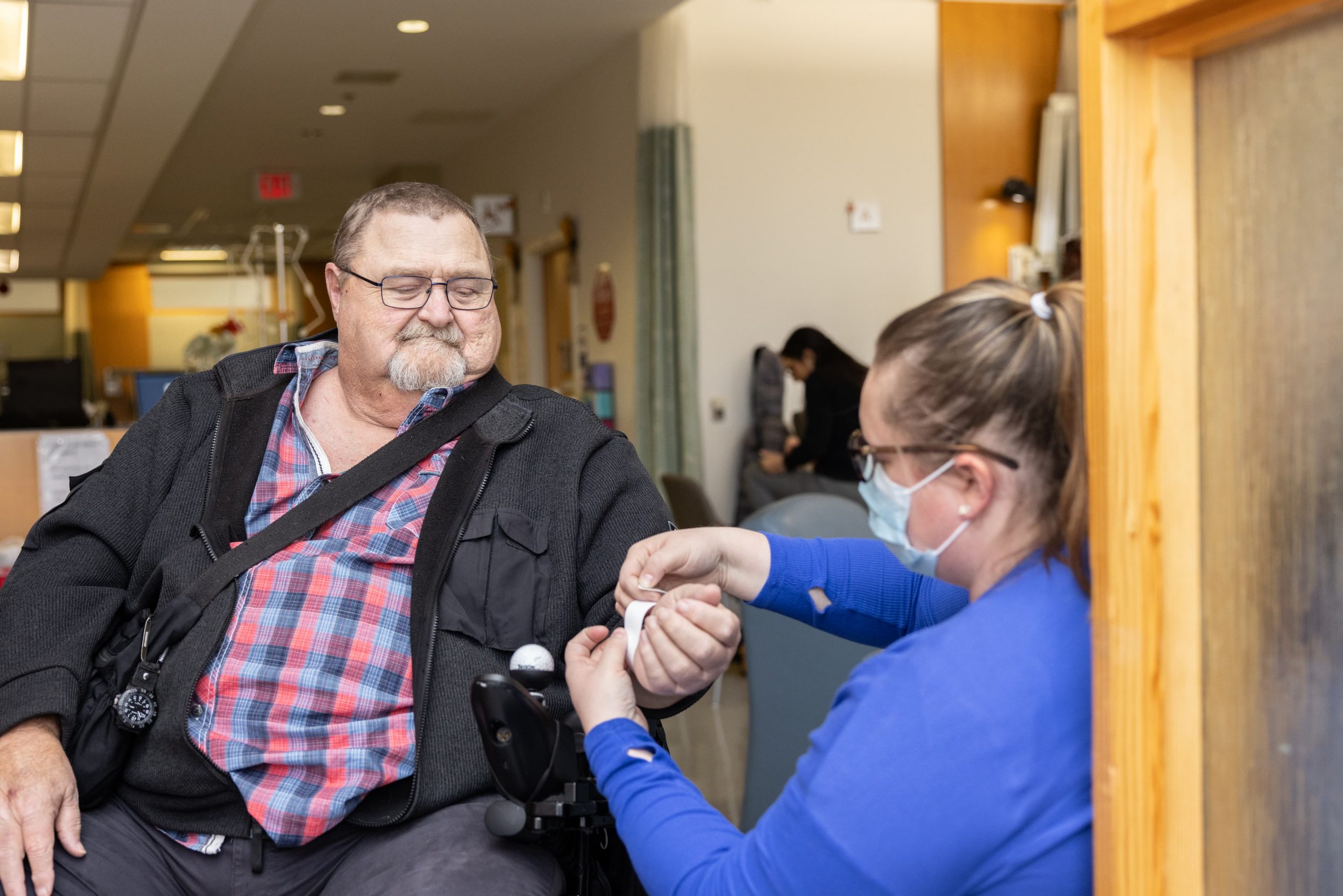 A healthcare worker in a mask assists a seated man with a medical device on his arm in a clinical setting.