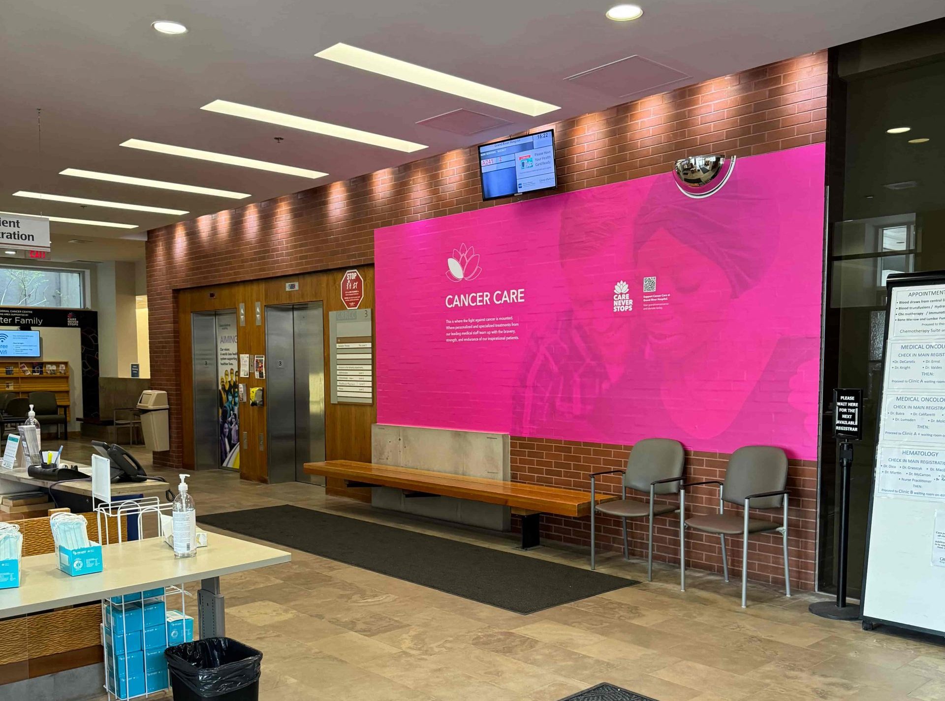 Hospital lobby with a pink Cancer Care information wall, chairs, benches, and a reception desk in the foreground.