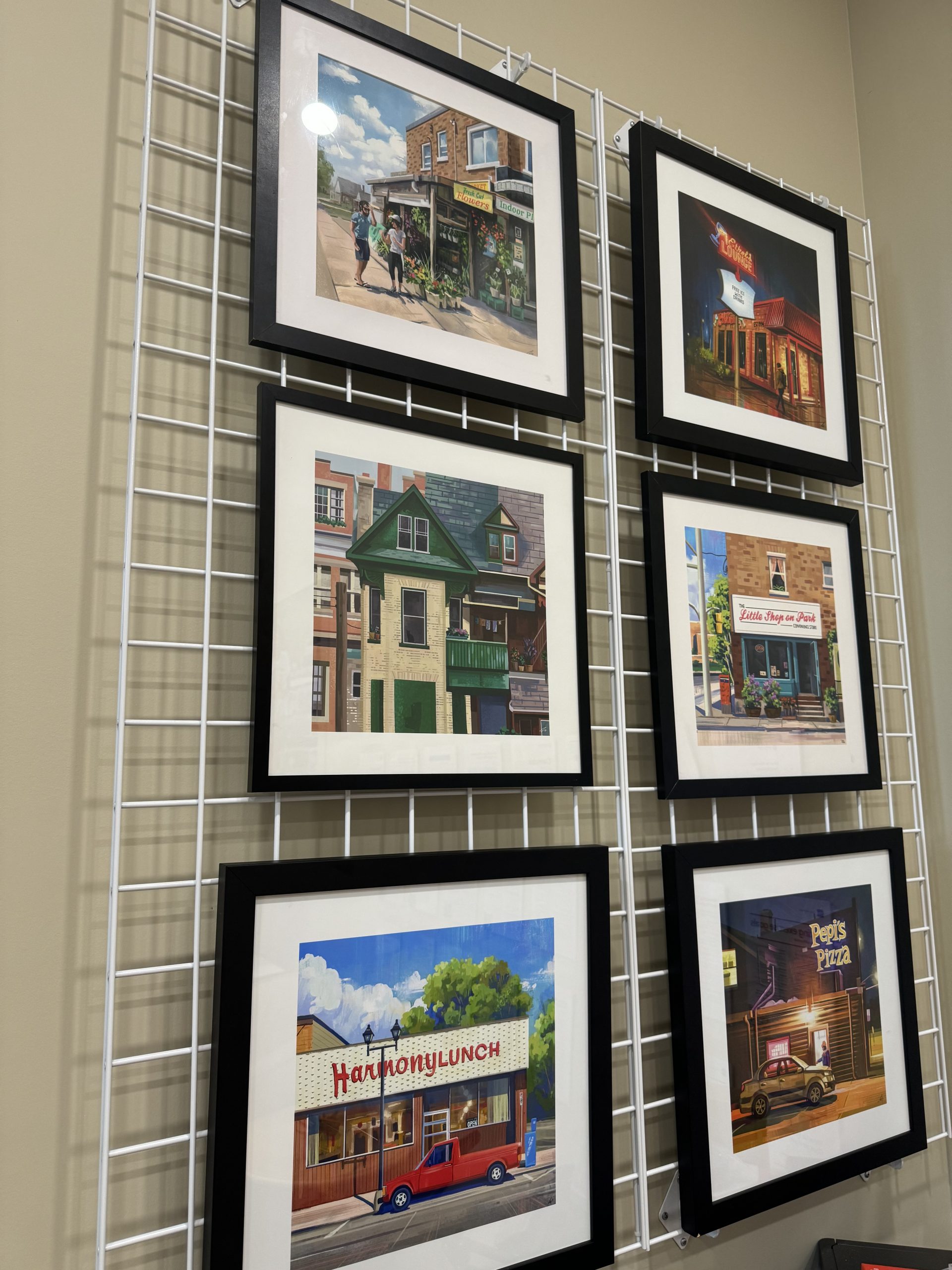 Six framed paintings of various storefronts are displayed on a white metal grid against a beige wall.