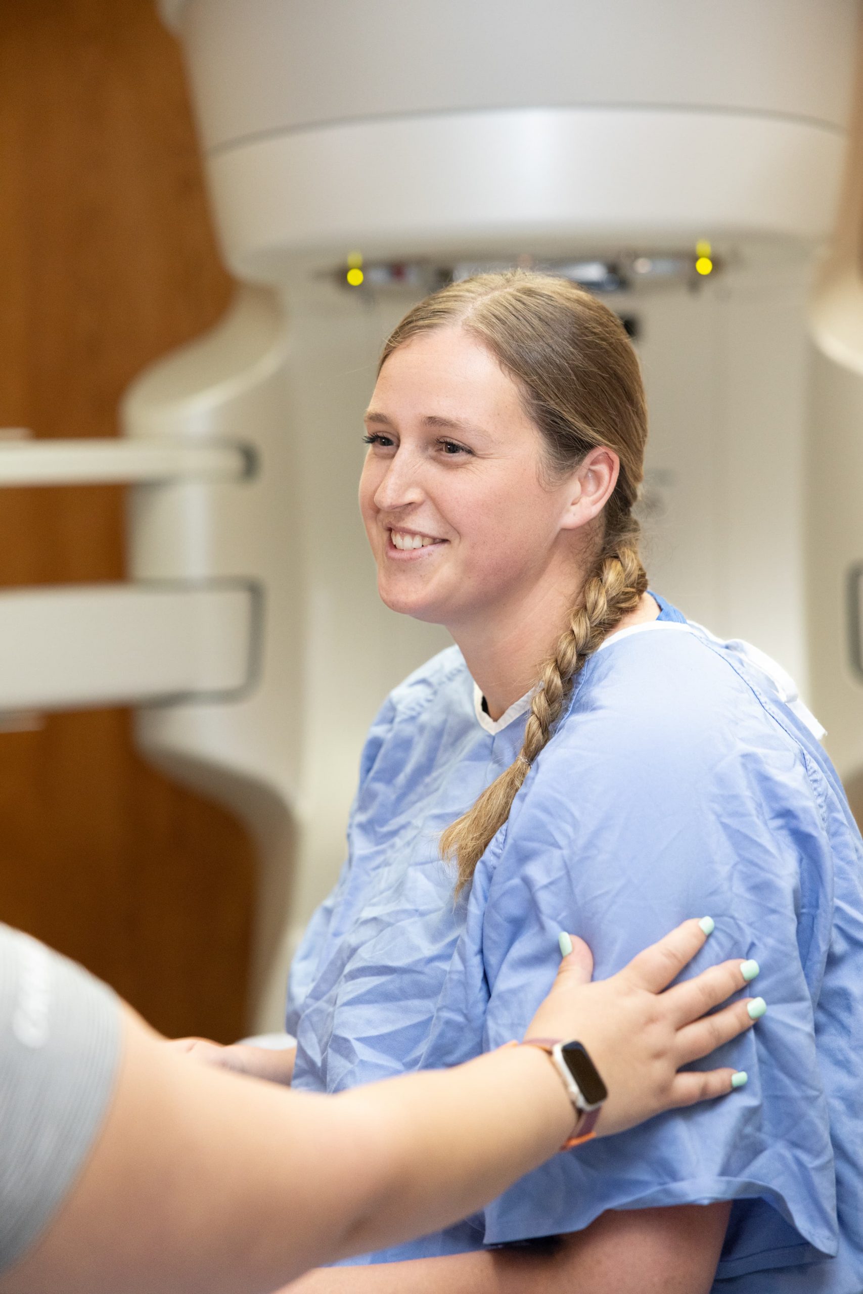 A patient in a hospital gown sits and smiles as another person touches their arm in a medical setting.