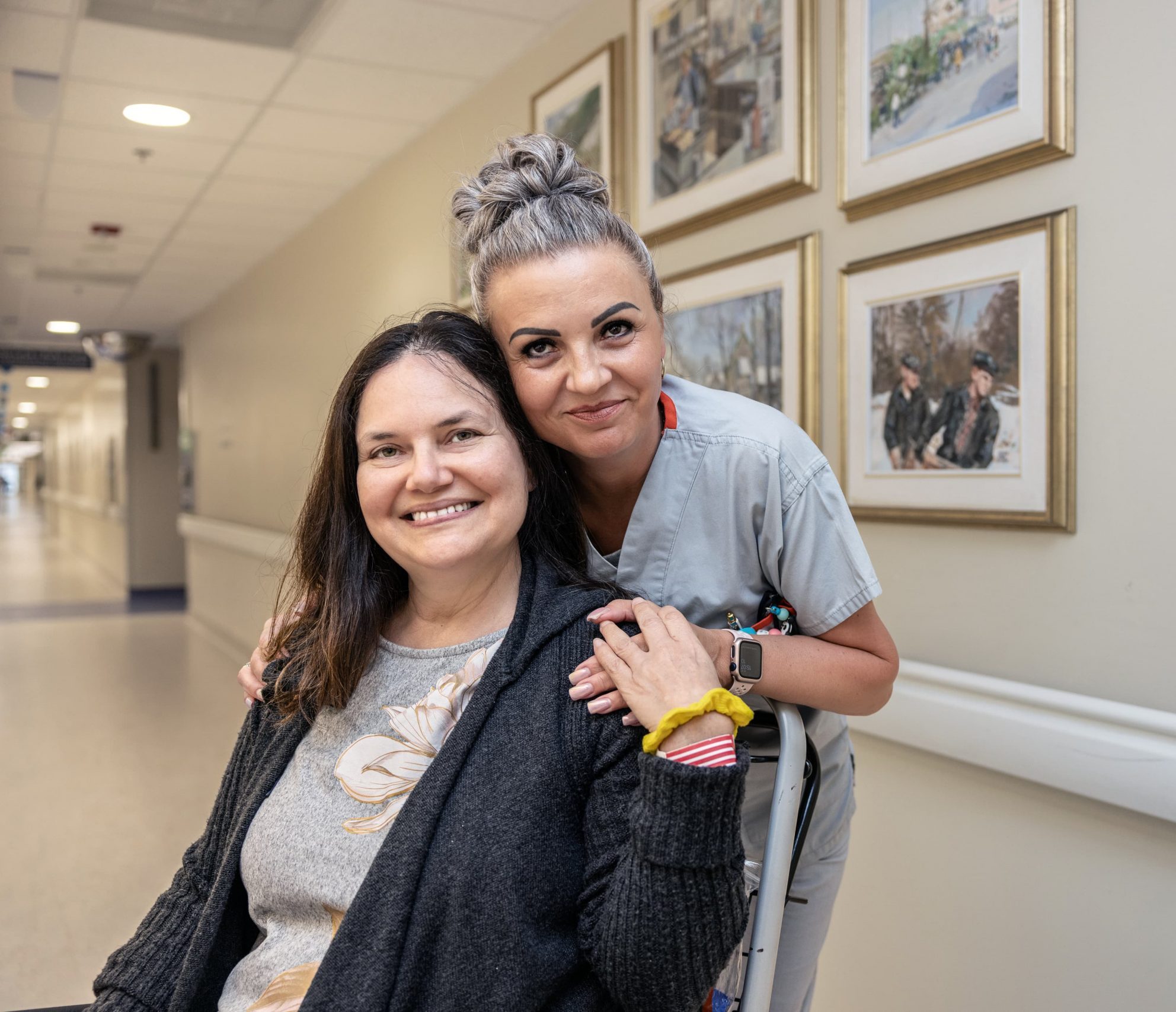 A smiling porter stands behind a seated patient in a hallway decorated with framed pictures.