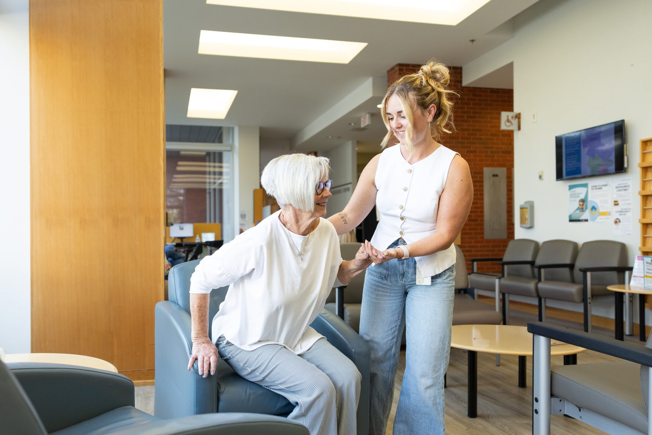 A young woman helps an older woman stand up from a chair in a brightly lit waiting area.