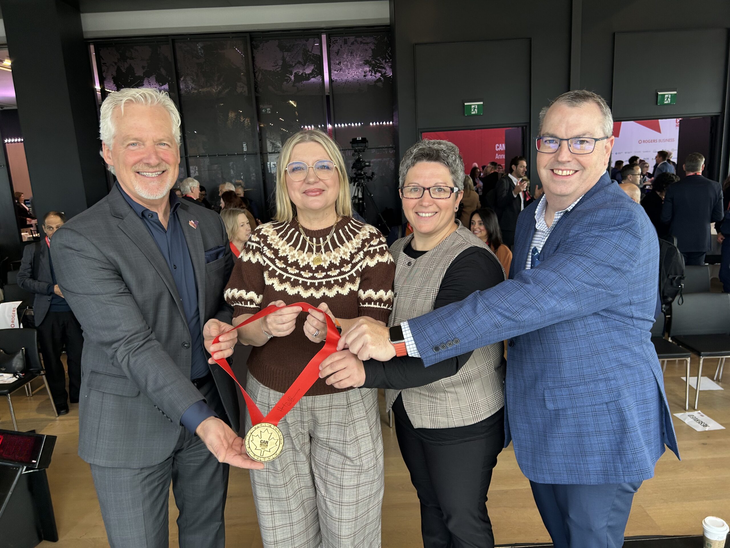 Quatre personnes debout à l'intérieur, tenant ensemble une grande médaille d'or, souriant à la caméra lors d'un événement.
