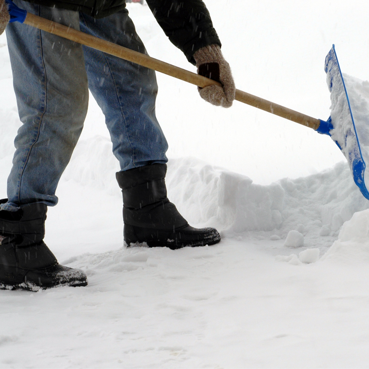 Une personne portant des bottes et des gants d'hiver utilise une pelle à neige bleue pour déblayer la neige du sol.