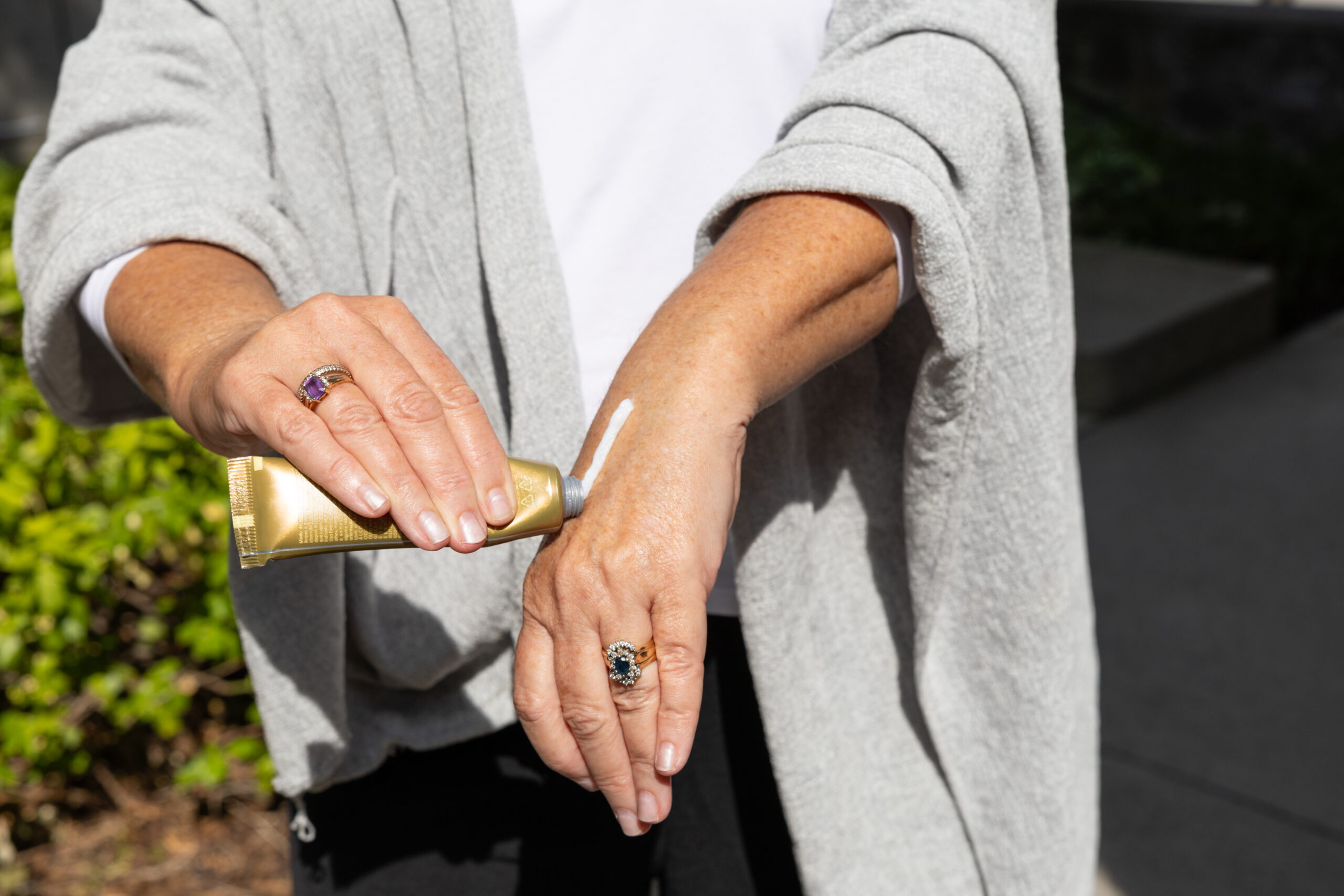 A person applies lotion from a gold tube onto the back of their hand outdoors, wearing a light gray cardigan.