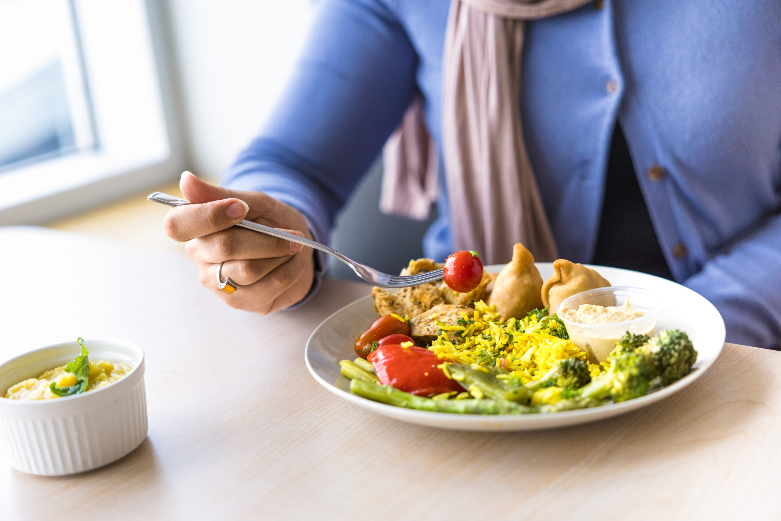 Person eating a plate of assorted vegetables, rice, and dumplings with a fork, with a small bowl of dip on the side.