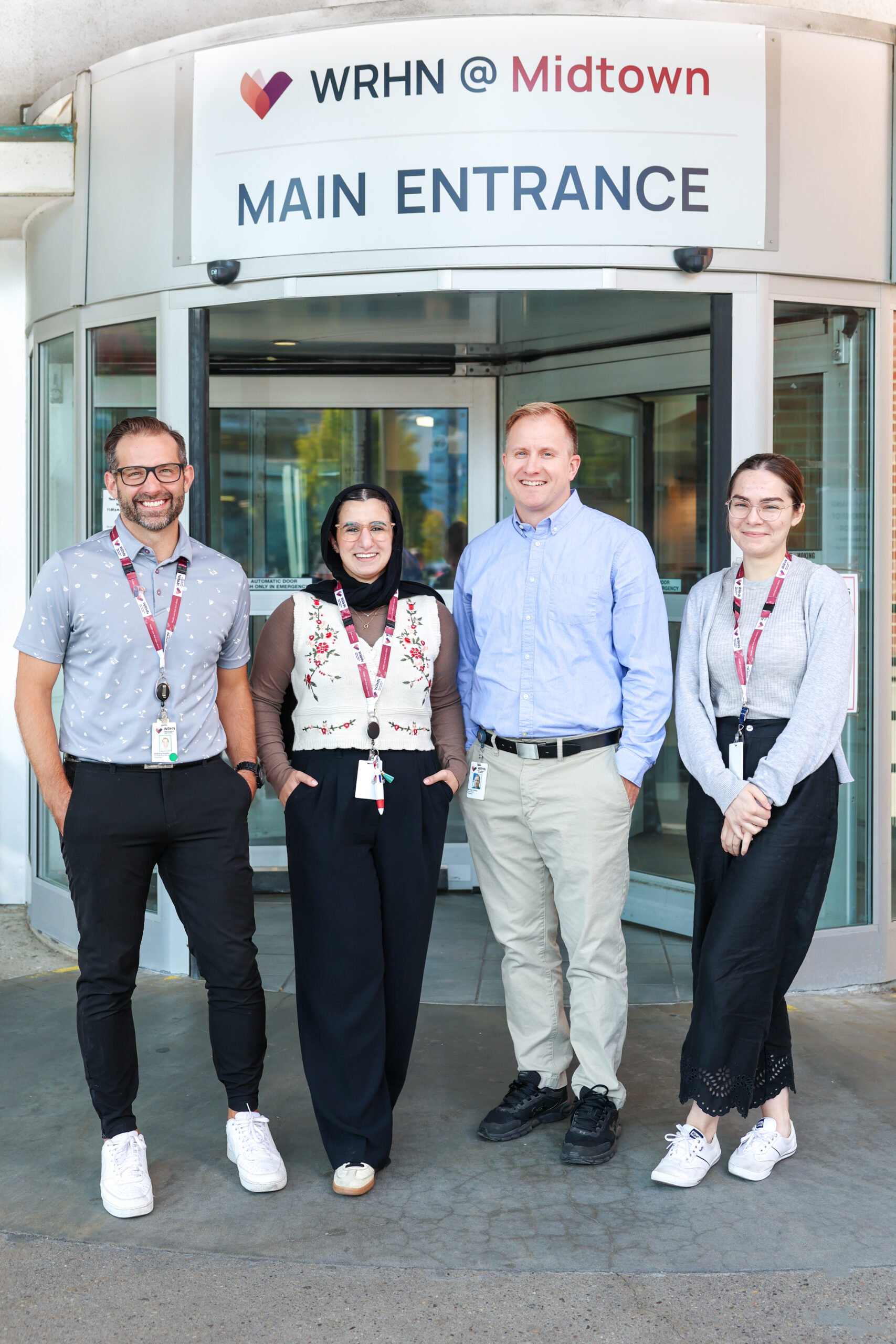 Quatre personnes se tiennent debout et sourient devant l'entrée principale du WRHN @ Midtown, portant des badges nominatifs et des cordons.