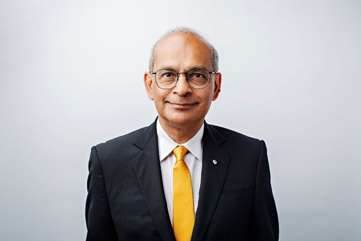 Retrato profesional de busto de un hombre con gafas, traje oscuro, camisa blanca y corbata de color amarillo brillante. Mira a la cámara con una leve sonrisa sobre un fondo gris claro. La imagen tiene un aspecto limpio y formal, ideal para un perfil profesional o una biografía corporativa.
