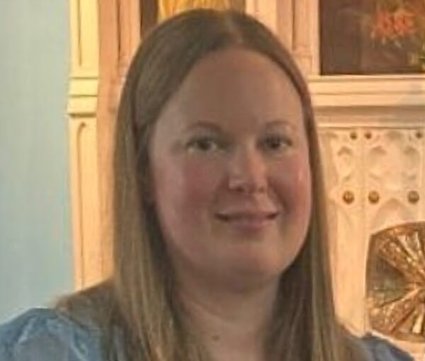 Woman with long brown hair wearing a light blue blouse, smiling at the camera indoors.